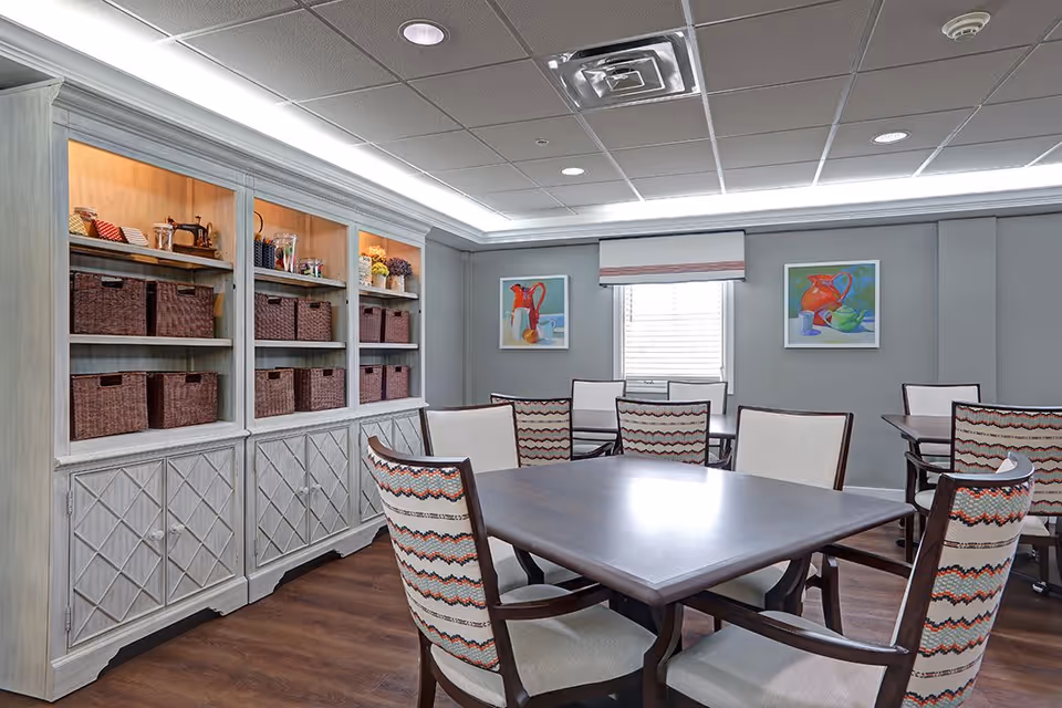 A well-lit room with multiple tables and chairs arranged for group seating. The chairs have patterned upholstery and the tables have a dark wood finish. Along one wall, there is a large built-in shelving unit with baskets and decorative items. Two colorful paintings hang on the opposite wall, and a window with a blind is centered between them. The ceiling has recessed lighting and a drop ceiling design.