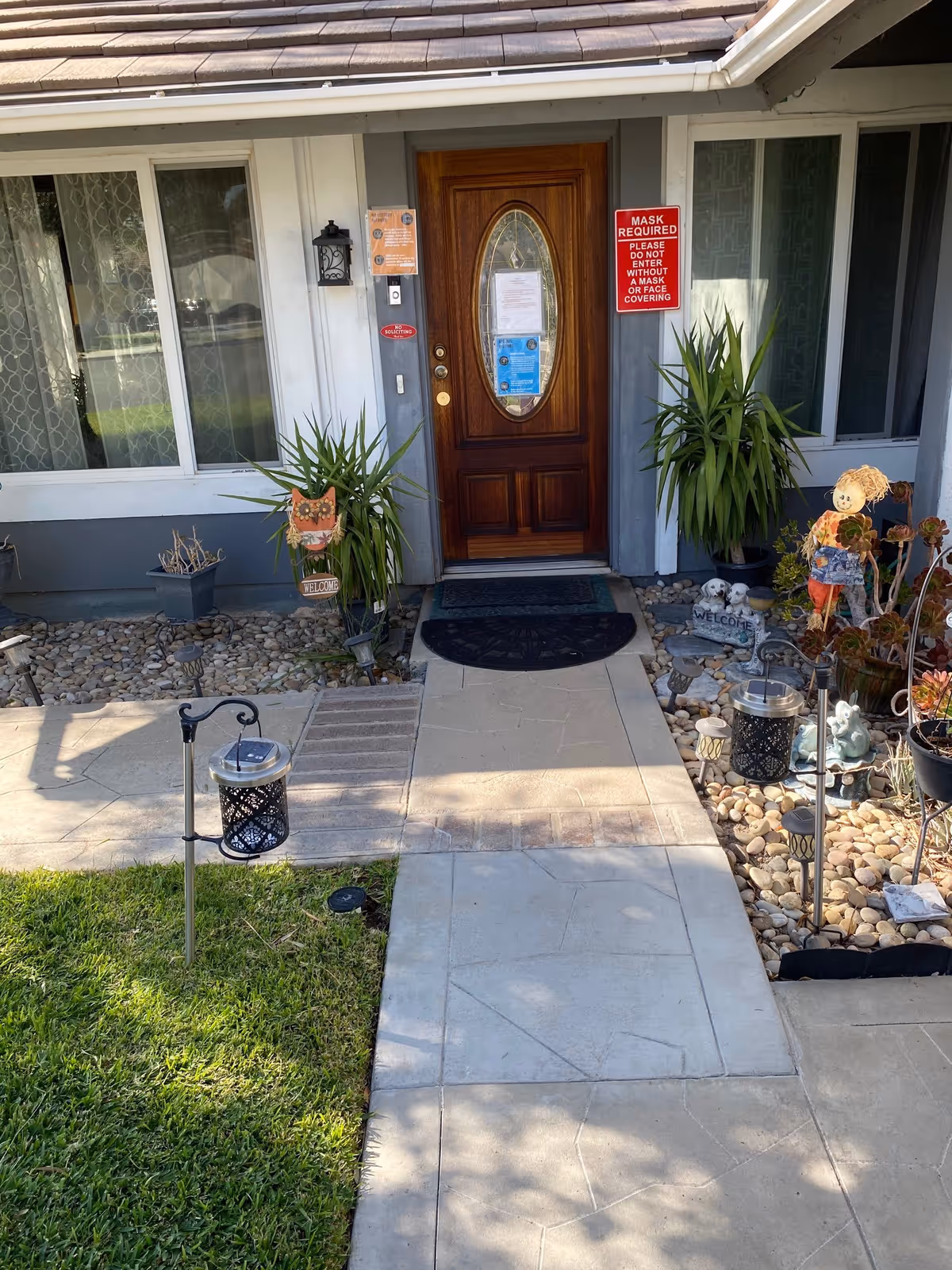 Front entrance of a residential building with a wooden door featuring an oval glass panel. There are potted plants on either side of the door, decorative garden ornaments including a small scarecrow figure and welcome signs, and a pathway leading up to the door. A red sign on the wall next to the door reads 'MASK REQUIRED PLEASE DO NOT ENTER WITHOUT A MASK OR FACE COVERING.'