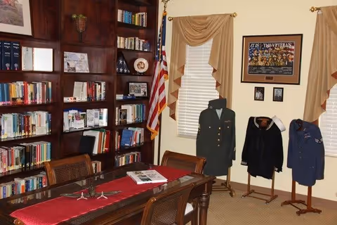 A room with wooden bookshelves filled with books and decorative items, an American flag, and three mannequins displaying military uniforms. There is a wooden table with a red runner and some papers on it. The walls have framed pictures and windows with beige curtains.