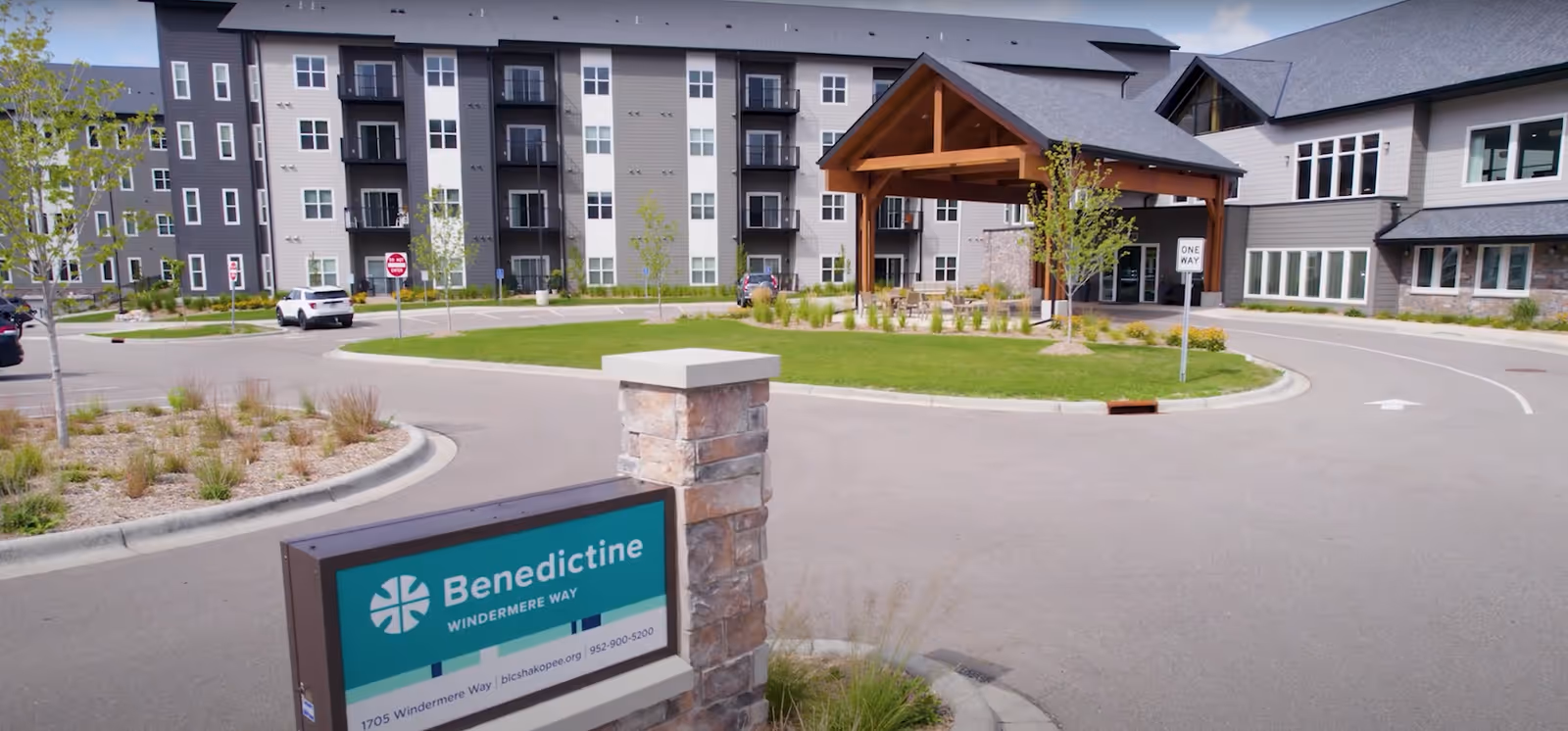 Exterior view of Benedictine Living Community - Shakopee Windermere Way, showing a multi-story building with balconies, a covered entrance with wooden beams, a circular driveway, landscaped greenery, and a sign with the facility's name and contact information.