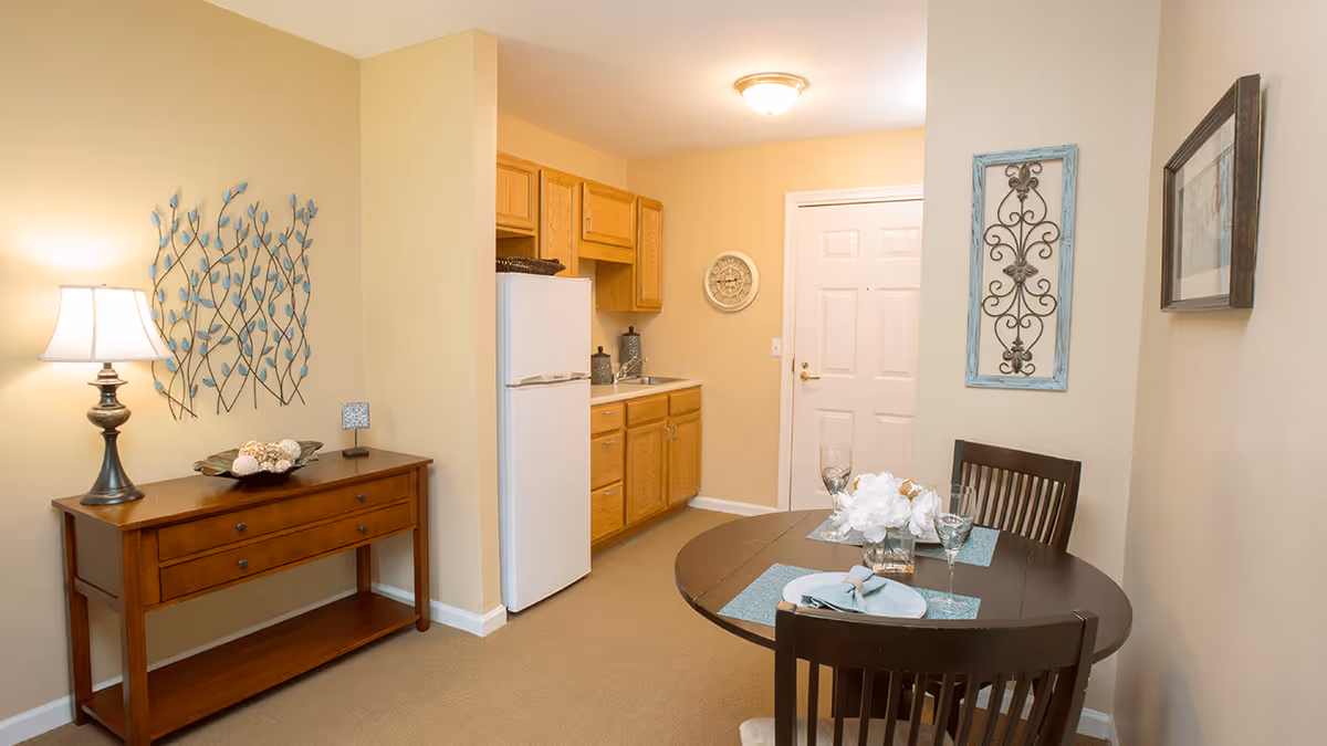 A small dining area and kitchenette in a senior living facility. The dining area features a round dark wooden table set for two with plates, glasses, and a white floral centerpiece. The kitchenette has wooden cabinets, a white refrigerator, and a sink. A wooden console table with a lamp and decorative items is against the wall, which is adorned with metal and framed wall art. The walls are painted beige and the floor is carpeted.