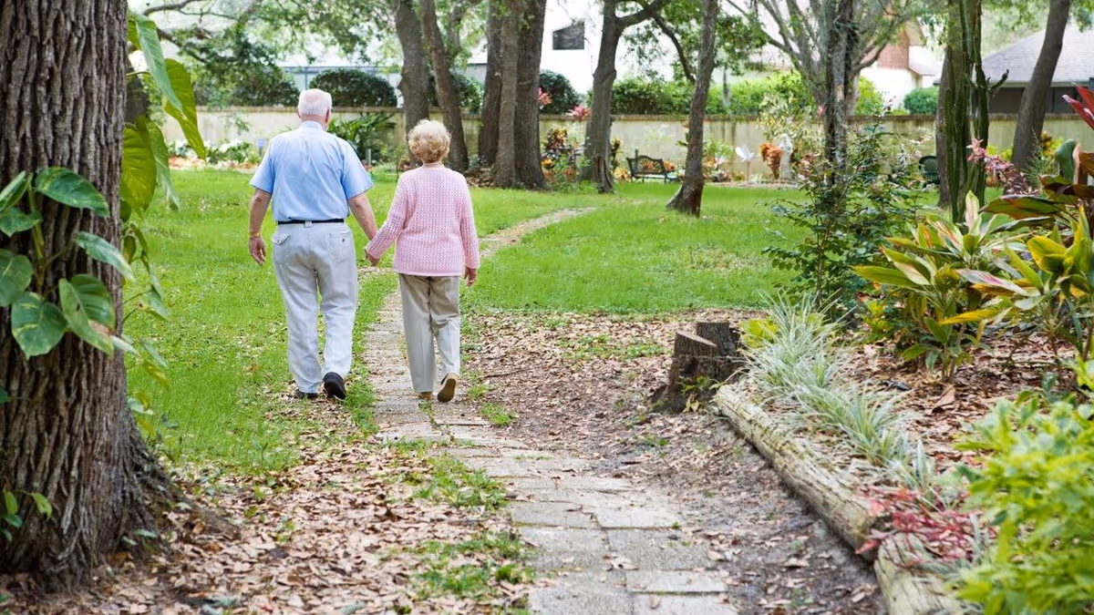 An elderly couple walking hand in hand down a paved garden path surrounded by trees, grass, and various plants in a peaceful outdoor setting.