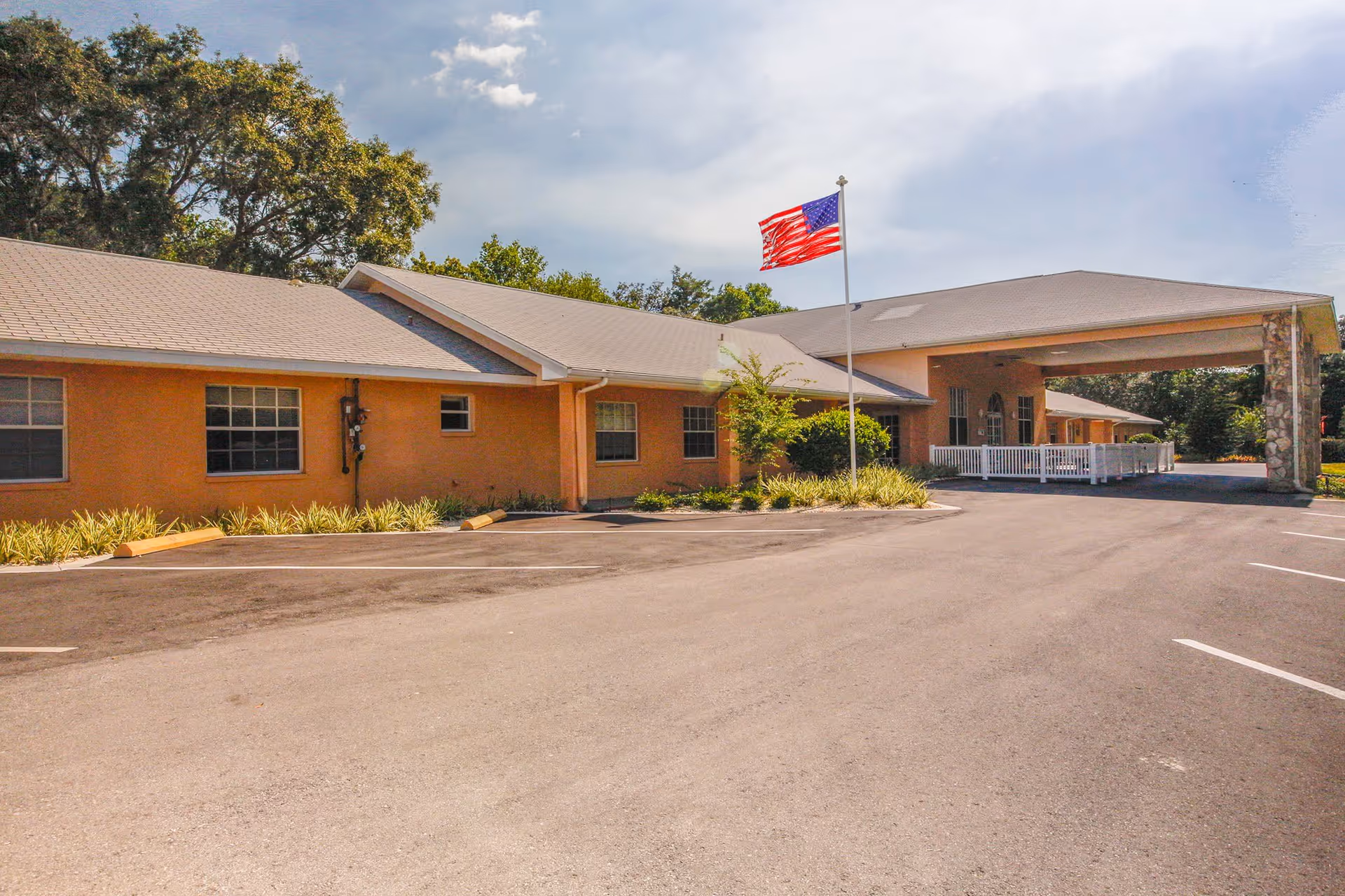 Exterior view of Jennifer Gardens Assisted Living and Memory Care building with a large covered entrance, an American flag on a flagpole, and a parking lot in front. The building is surrounded by trees and landscaping.