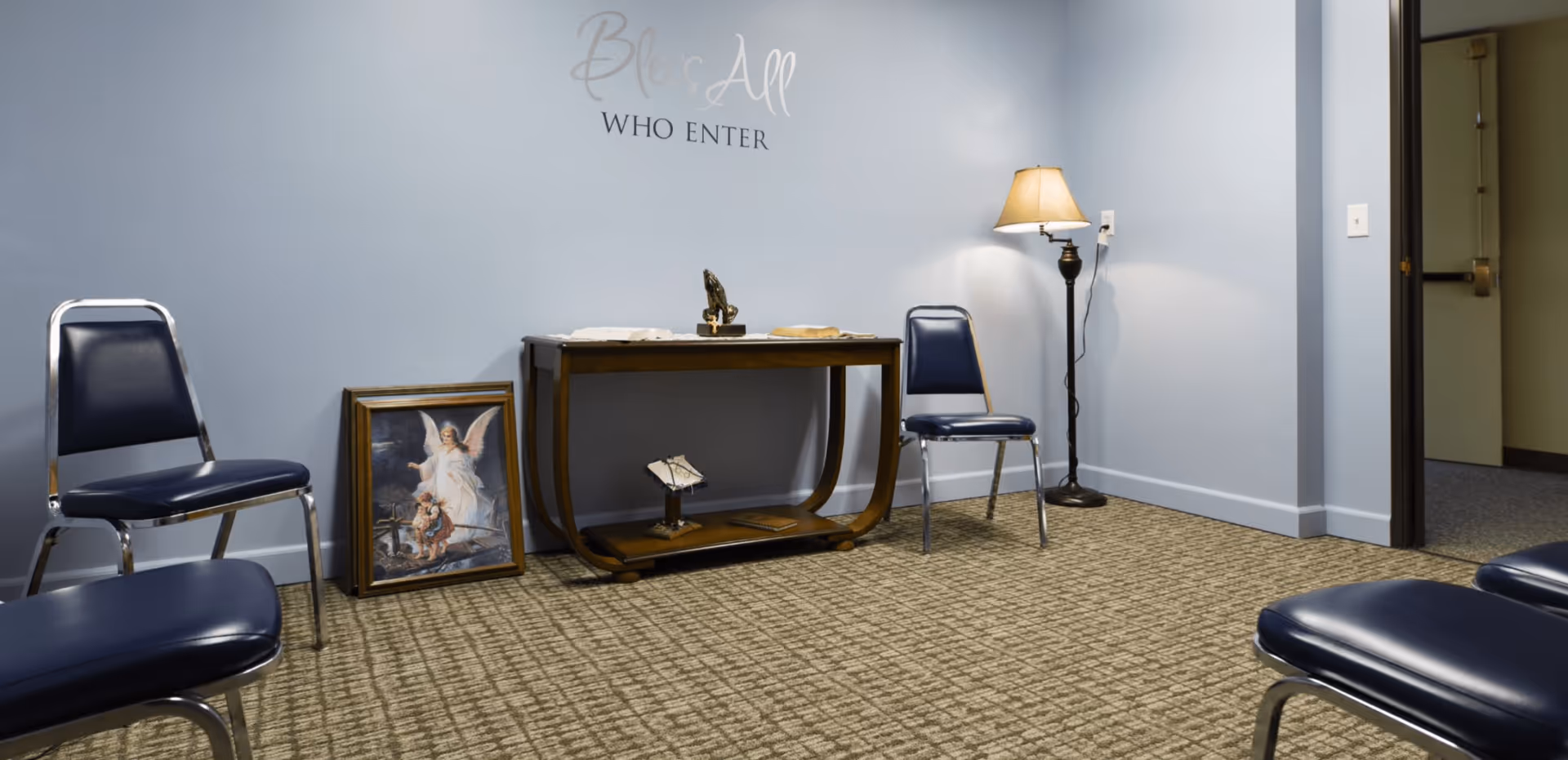 A small room with light blue walls and beige carpet featuring four blue padded chairs arranged around the edges. A wooden table against the wall holds religious items including a praying hands statue and a book. A framed picture of an angel with two children leans against the wall. A floor lamp with a beige shade is next to one chair. Above the table, a wall decal reads 'Bless All Who Enter'. An open door leads to another room.