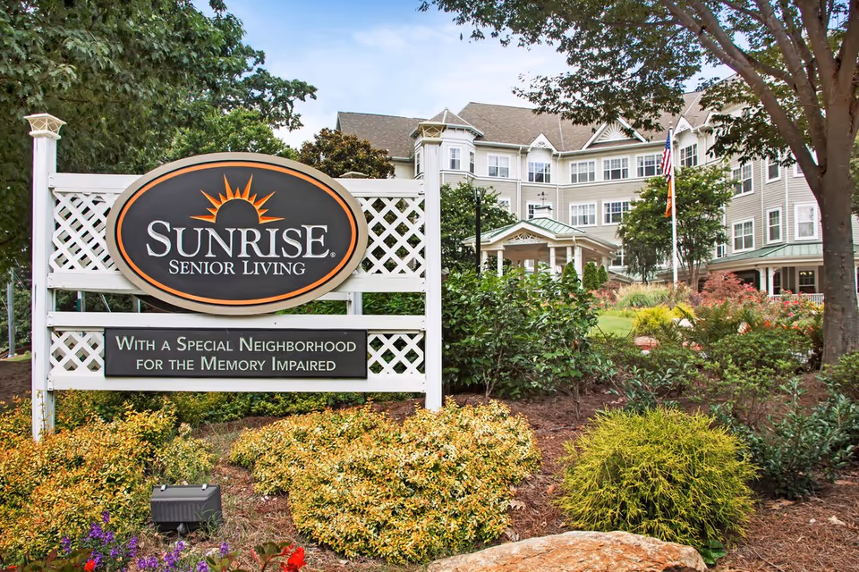 Outdoor view of Sunrise Senior Living facility with a large sign in the foreground that reads 'Sunrise Senior Living' and 'With a Special Neighborhood for the Memory Impaired'. The background shows a multi-story building surrounded by trees, bushes, and landscaped gardens under a partly cloudy sky.