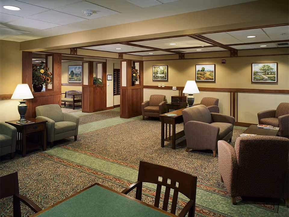 A spacious, well-lit lounge with upholstered chairs, side tables, lamps, and framed artwork in a senior living facility.