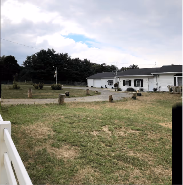 Single-story white building with a grassy yard, gravel driveway, tree stumps, and a flagpole under a cloudy sky.