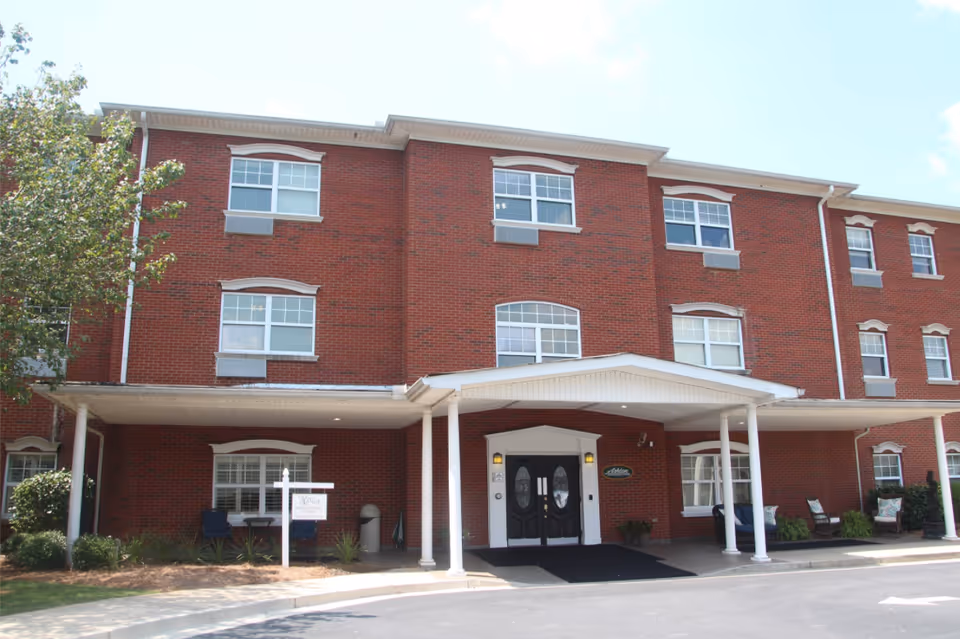 Front exterior view of a three-story red brick building with white trim and multiple windows. The entrance has a covered porch supported by white columns, with double black doors and a sign that reads 'Ashton Manor'. There are chairs and plants near the entrance and a tree on the left side.