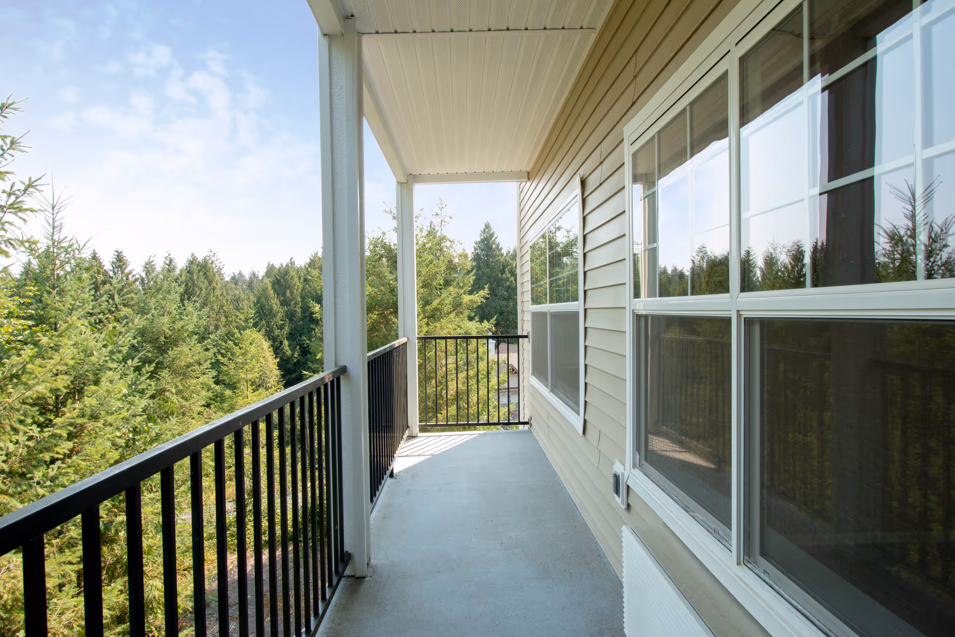 A covered balcony with black metal railings overlooking a lush green forest. The balcony has a light gray floor and beige siding on the building wall with large windows reflecting the trees outside.