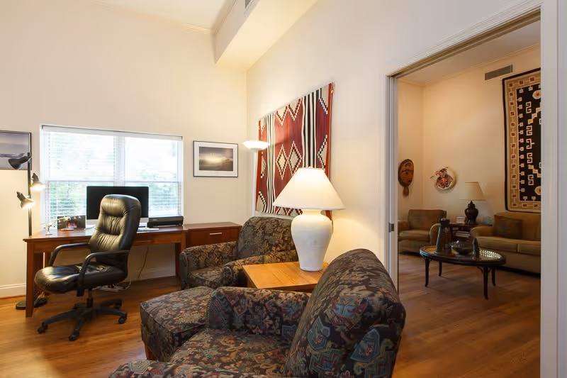 A cozy interior room with two patterned armchairs and a wooden side table with a white lamp. In the background, there is a wooden desk with a black office chair and a computer monitor in front of a window with blinds. The walls are decorated with framed pictures and a large textile wall hanging with geometric patterns. Through an open doorway, another room is visible with a sofa, armchairs, a round coffee table, and additional wall decorations.
