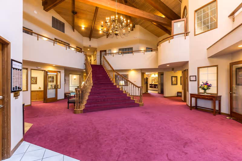 Spacious senior living facility lobby with a large red carpeted staircase leading to an upper floor. The area features wooden railings, a chandelier hanging from a high wooden ceiling with exposed beams, and several doorways and windows. A small table with a flower arrangement is visible on the right side.