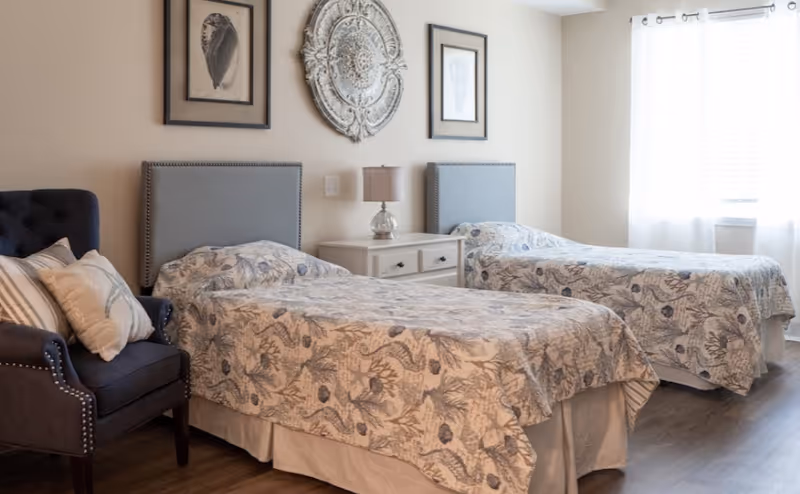 A bright bedroom with two twin beds featuring patterned bedspreads, a white nightstand with a lamp between the beds, a dark upholstered armchair with pillows on the left, and three decorative wall hangings above the beds. A window with white curtains allows natural light into the room.