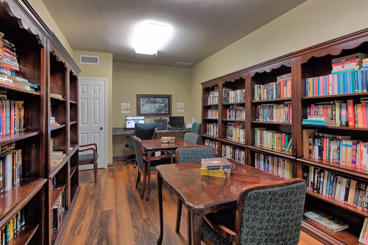 Interior view of a library room at Town Village Tulsa with wooden bookshelves filled with books on both sides. There are two wooden tables with green upholstered chairs in the center. At the back of the room, there is a desk with two computer monitors and office chairs, and a framed picture on the wall above the desk.