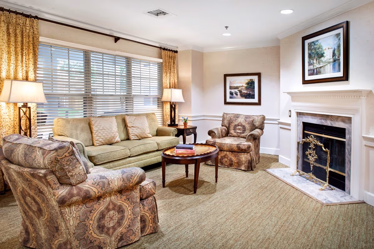A cozy living room with a beige sofa, two patterned armchairs, a round wooden coffee table with books, two table lamps, large window with blinds and curtains, a fireplace with a decorative screen, and framed artwork on the walls.