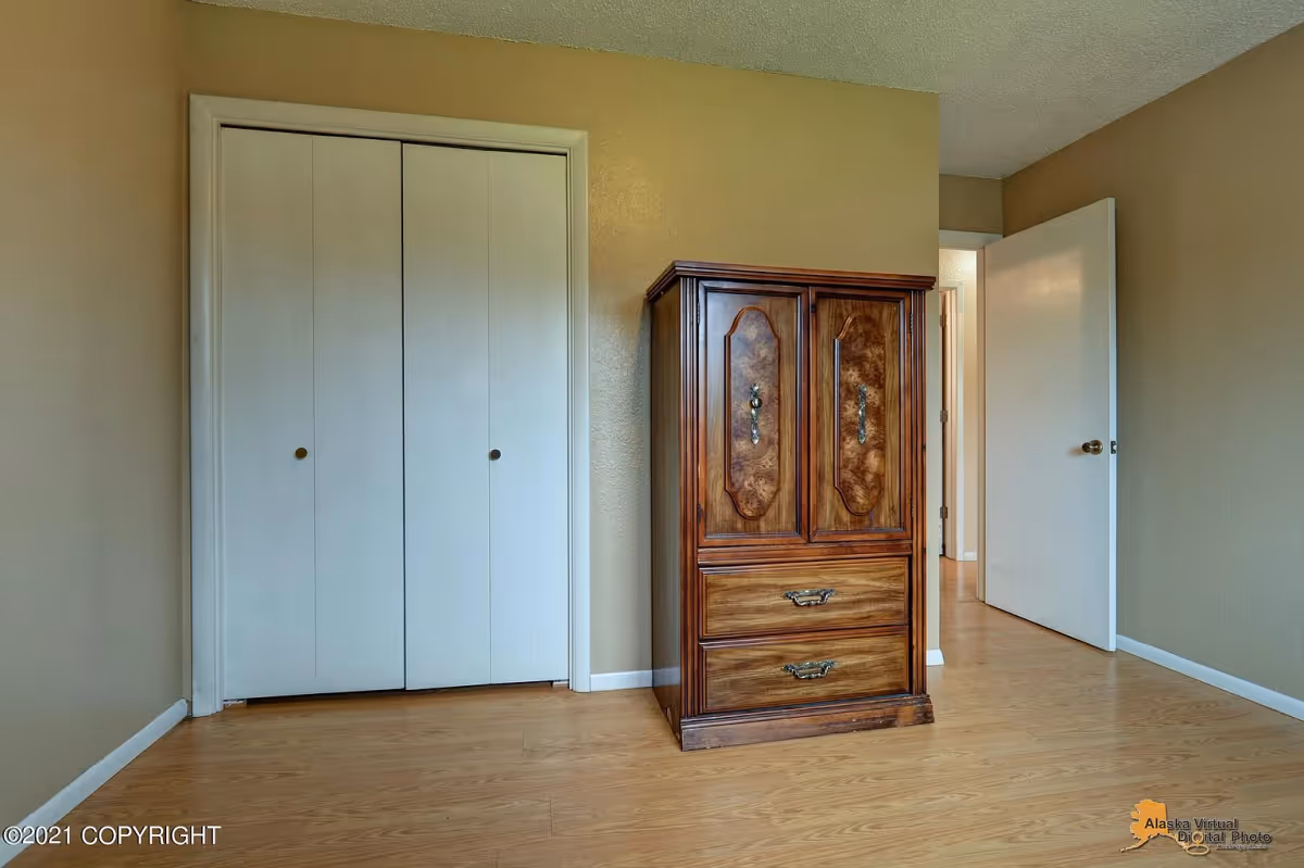 An empty room with beige walls and light wood flooring featuring a closed white double-door closet on the left and a wooden cabinet with ornate handles on the right. An open white door leads to a hallway in the background.
