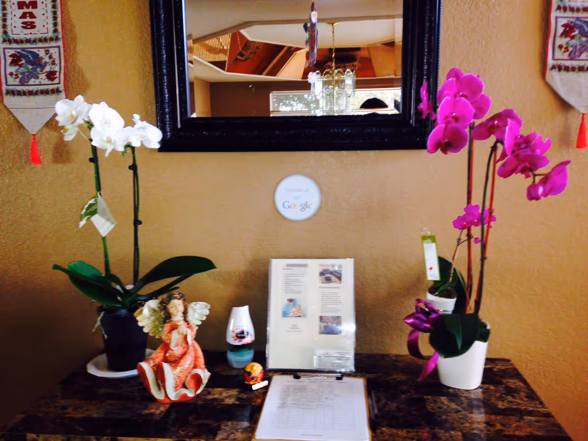 Entry table beneath a mirror displaying two orchid plants, an angel figurine, a clipboard sign-in sheet, and informational materials.