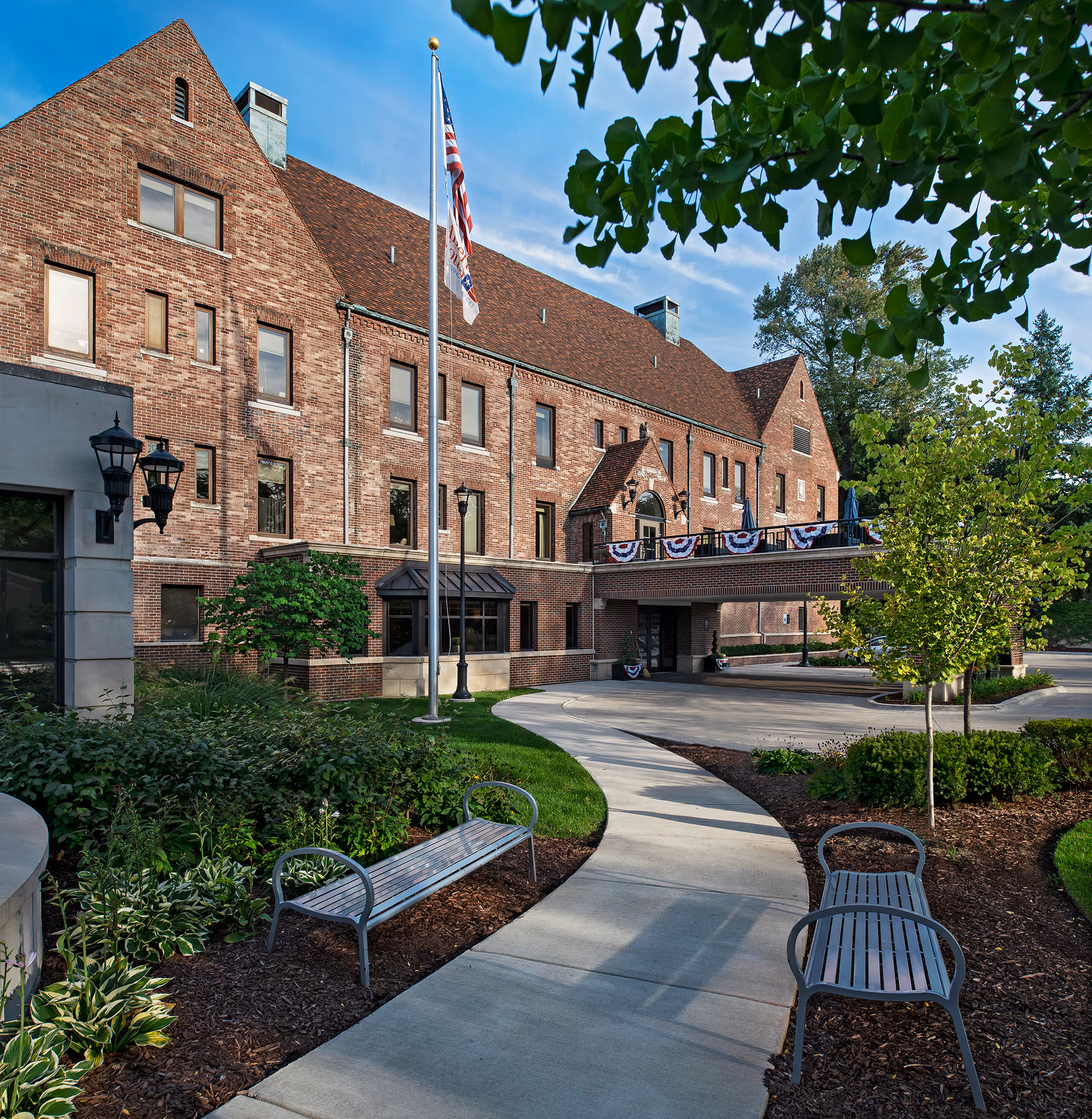 Exterior view of a large brick building with a peaked roof, an American flag on a flagpole, a curved concrete walkway, two metal benches, and landscaped greenery including trees and bushes under a blue sky.