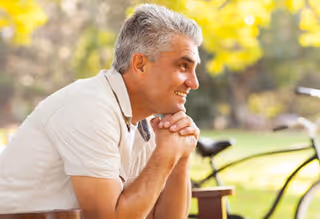 A smiling elderly man with gray hair wearing a light-colored polo shirt is sitting outdoors on a bench with his hands clasped, looking to the side. In the background, there is a blurred bicycle and green foliage with sunlight filtering through the trees.