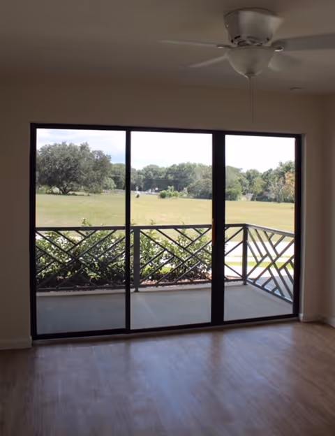 Empty room with wooden flooring and a ceiling fan, featuring large sliding glass doors that open to a balcony with a decorative railing and a view of a grassy field and trees outside.