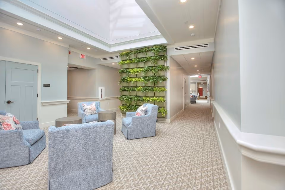 Bright interior lounge with upholstered chairs around low tables under a skylight and a vertical living wall beside a long corridor.