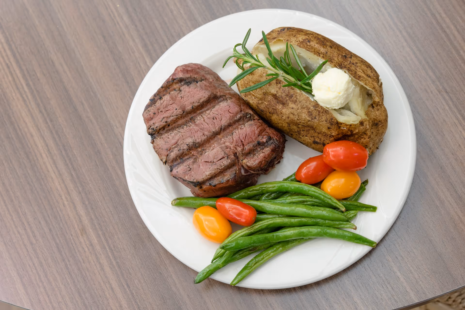 A white plate with a grilled steak, a baked potato topped with a dollop of butter and a sprig of rosemary, green beans, and red and yellow grape tomatoes on a wooden table.