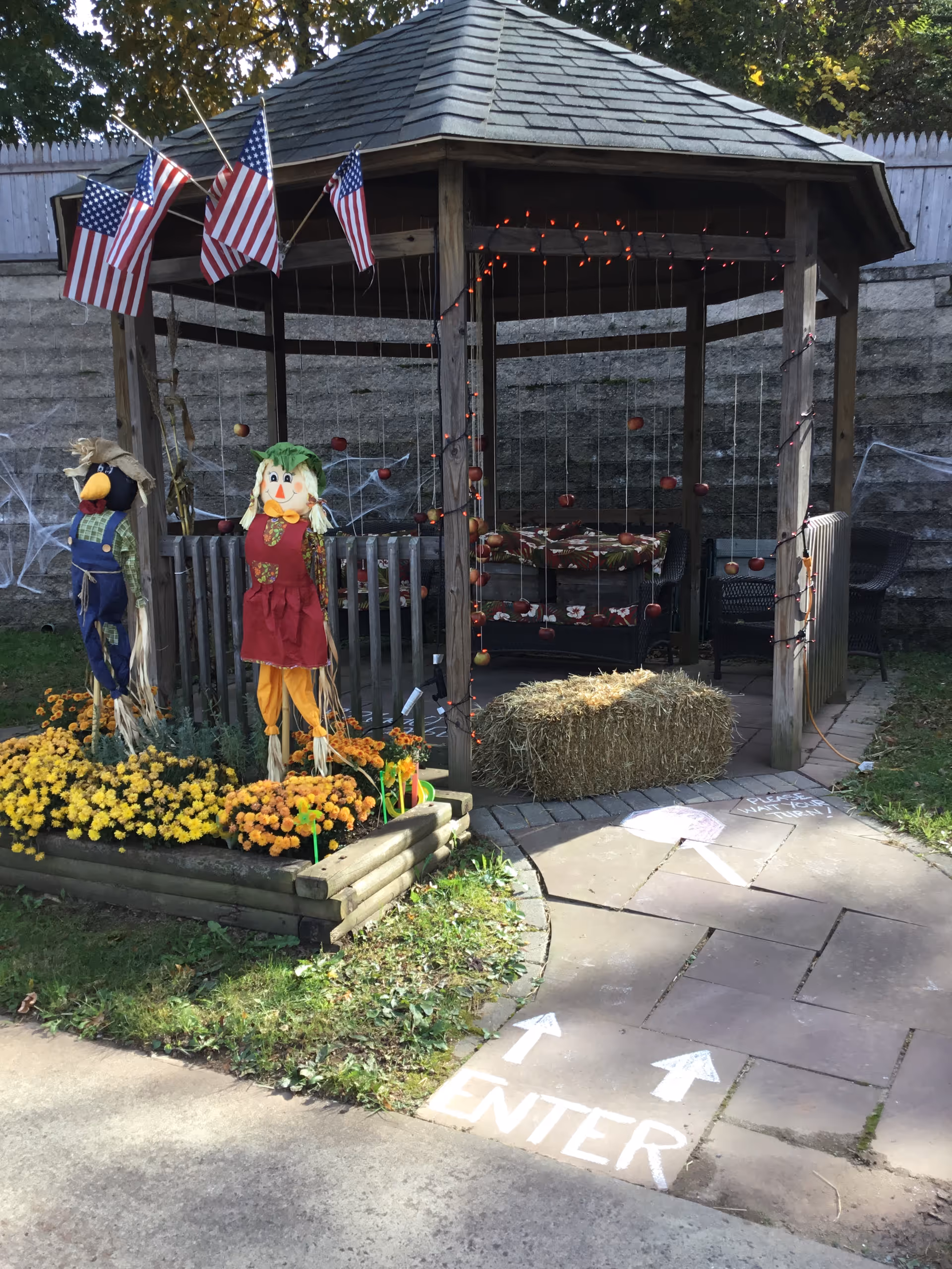 A wooden gazebo decorated with small American flags, orange string lights, and hanging red ornaments. In front of the gazebo, there are two scarecrows standing among yellow and orange flowers in a raised wooden flower bed. A bale of hay is placed on the stone-paved path leading to the gazebo. The path has white chalk markings with arrows and the word 'ENTER' directing visitors inside. The area is outdoors with grass and a stone wall in the background.