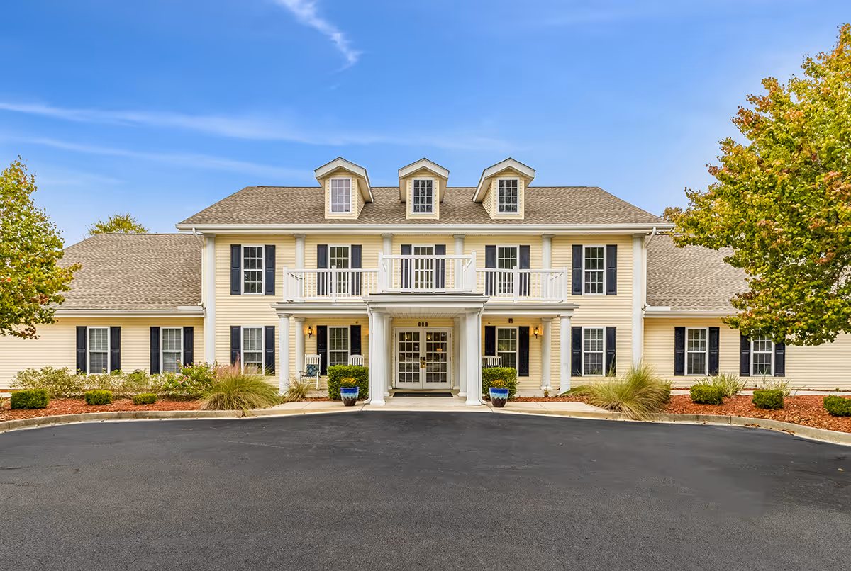 Front exterior view of a two-story senior living facility building with beige siding, black shutters, and a covered entrance with white columns. There are three dormer windows on the roof and landscaped bushes and trees on either side of the driveway.
