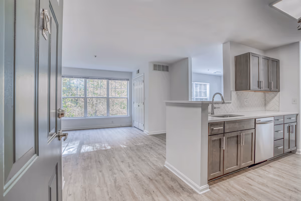 Open-plan apartment interior with a kitchen and island opening into a bright living area with large windows.