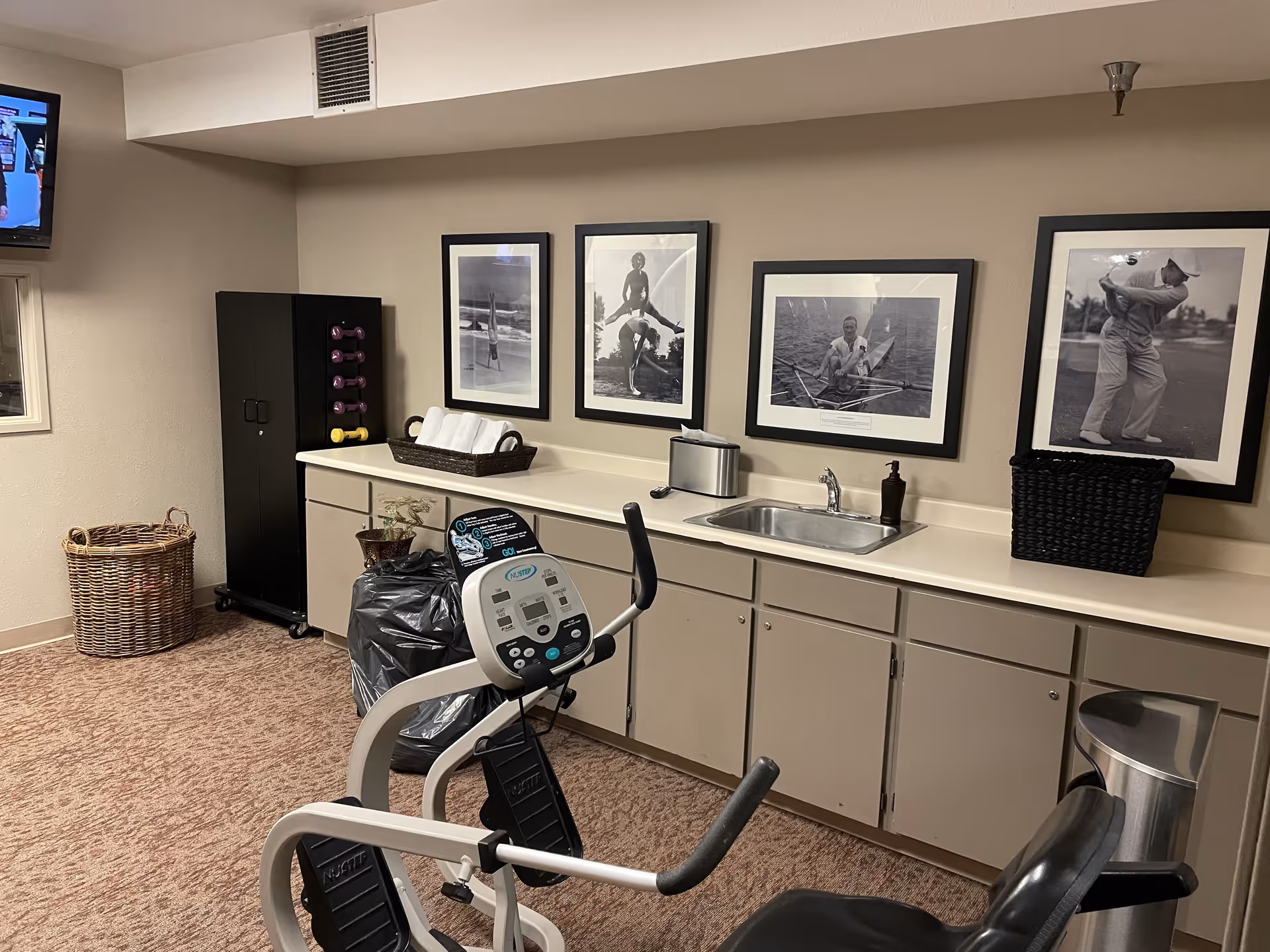 Interior room with exercise equipment including a stationary bike, a countertop with a sink, soap dispenser, and a basket. There are four black and white framed photos on the wall above the counter, a rack with small dumbbells, a wicker basket on the floor, and a television mounted on the wall.