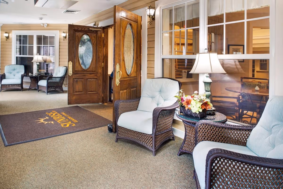A cozy indoor seating area with wicker chairs featuring light blue cushions, a small round wicker table with a floral arrangement and a lamp, and double wooden doors with oval glass panels leading to another room. A brown floor mat with the Sunrise logo is placed near the entrance.