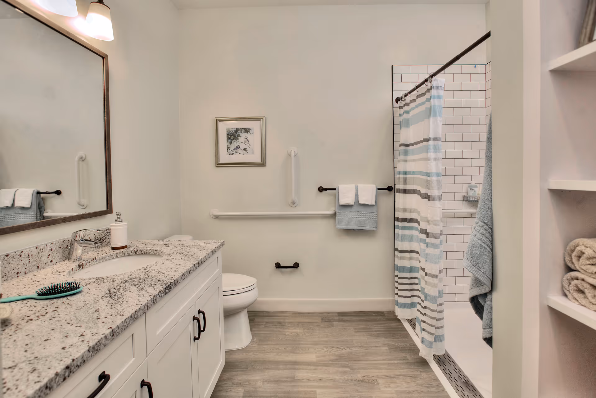 A clean and modern bathroom with a granite countertop vanity, a large mirror, a toilet, and a shower with a striped curtain. The bathroom has grab bars on the walls for accessibility, light wood flooring, and shelves with folded towels.