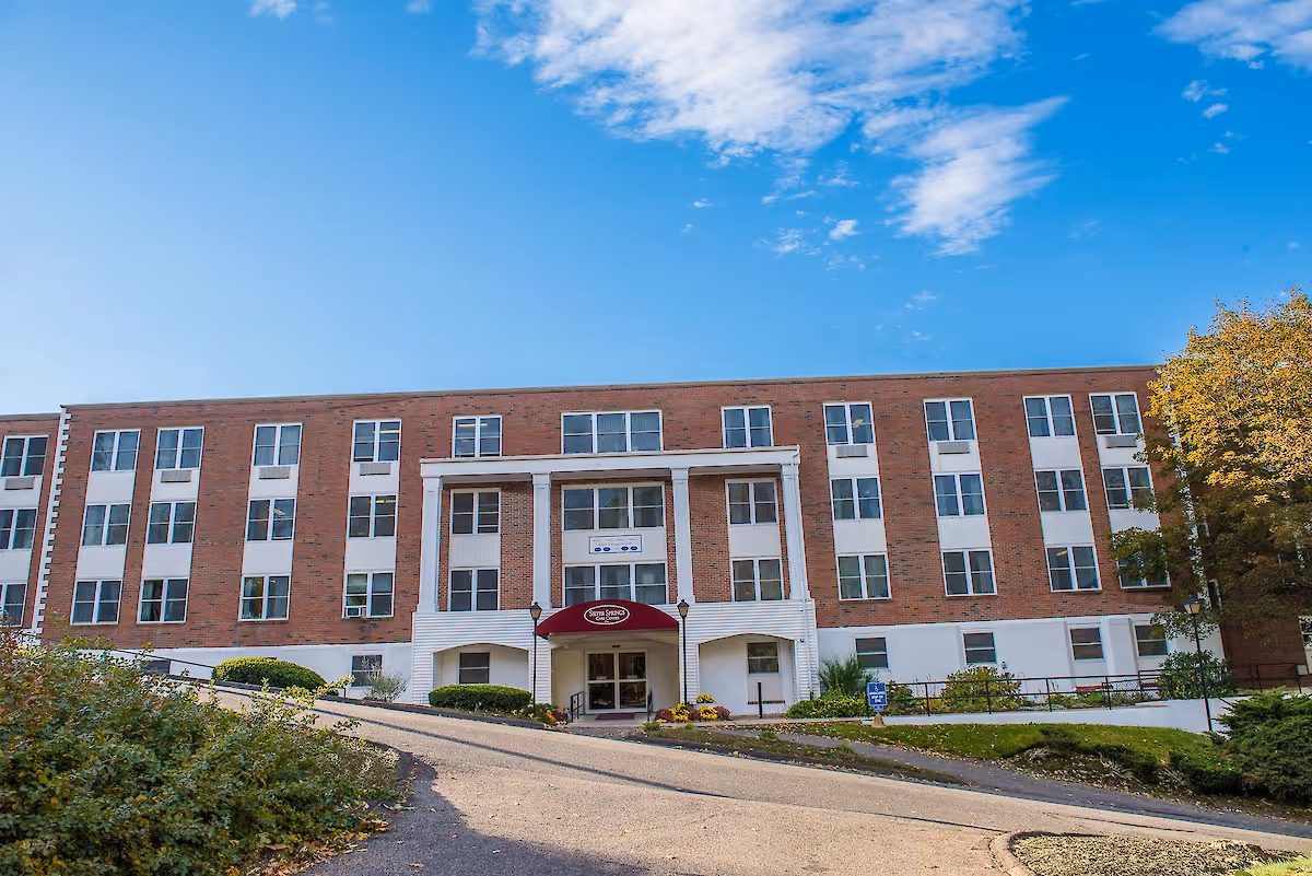 Front exterior view of a multi-story brick building with white accents and many windows under a blue sky with some clouds. The entrance has a red awning with the name Silver Springs Care Center. There is a paved driveway leading up to the entrance and some greenery around the building.