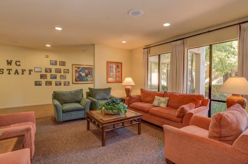 A cozy senior living common area with a red sofa, two red armchairs, two green armchairs, a wooden coffee table with a plant centerpiece, two table lamps, large windows with curtains letting in natural light, and framed pictures on the wall.