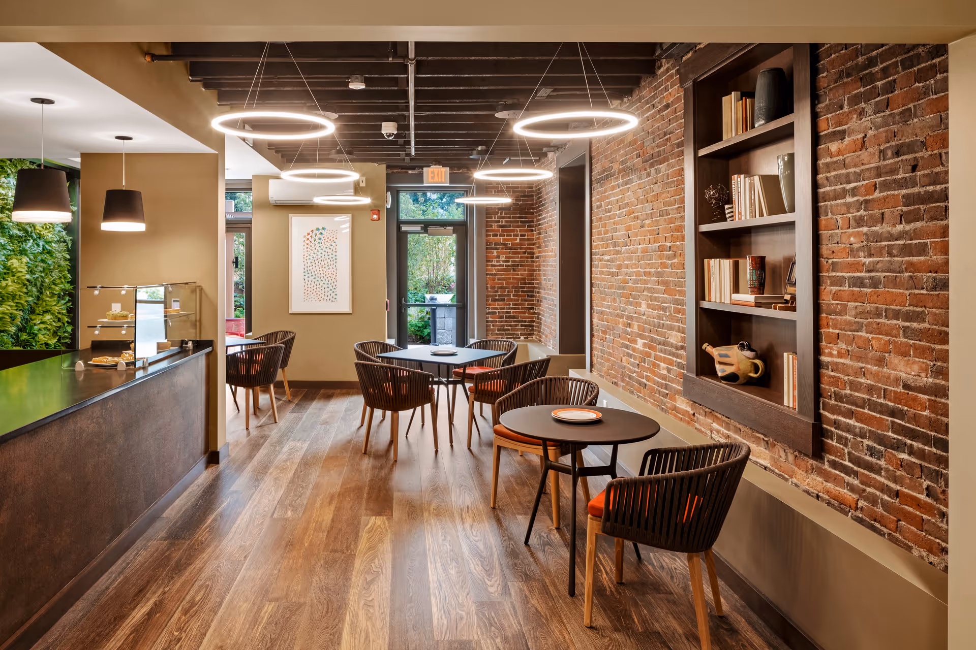 A modern dining area with round tables and chairs featuring orange cushions, exposed brick walls, wooden flooring, and circular hanging light fixtures. There is a built-in bookshelf on the right wall with books and decorative items. The left side has a counter with a glass display case and pendant lights above. A door and window at the far end show greenery outside.