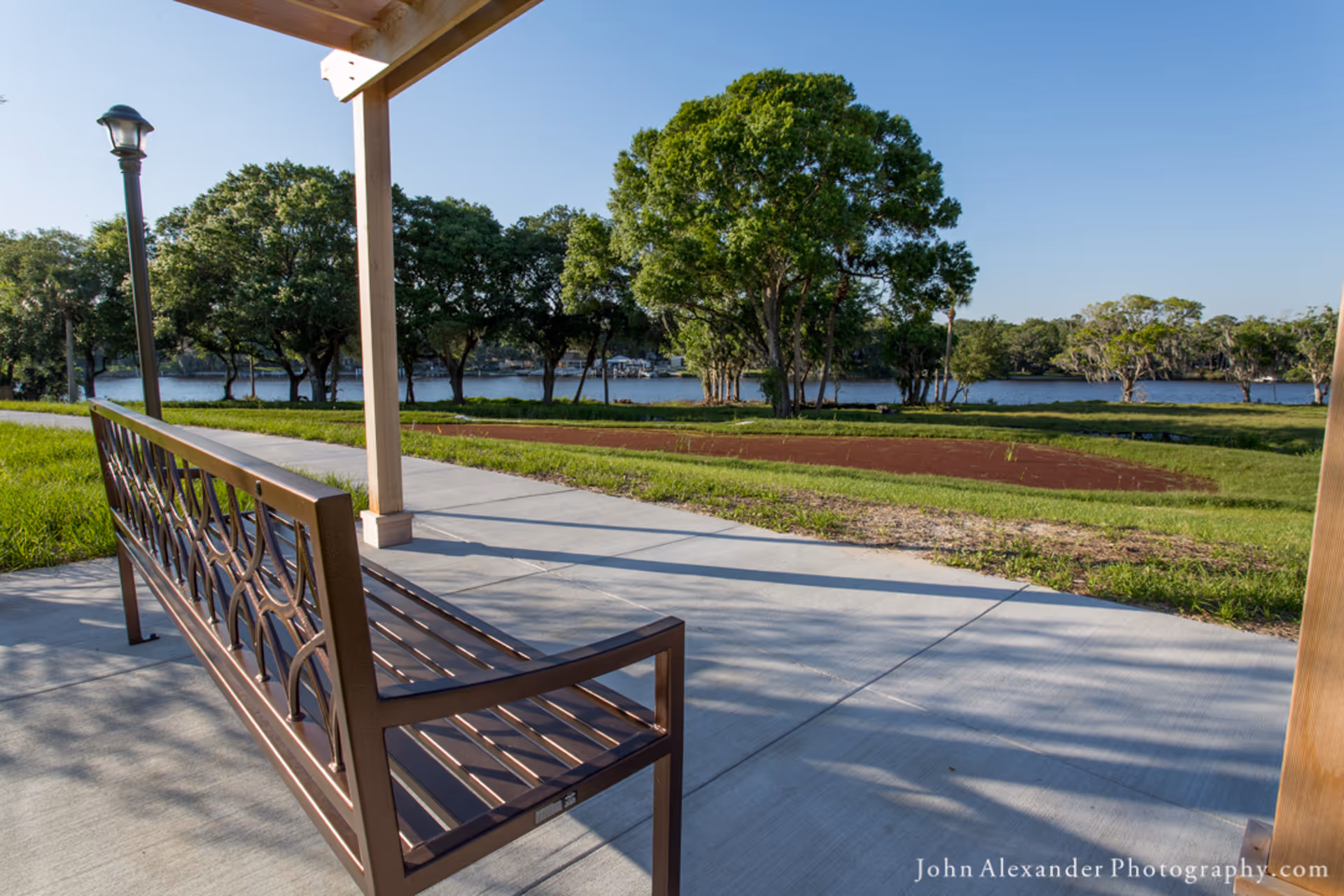 A metal bench under a wooden pergola on a concrete patio overlooking a grassy area with trees and a body of water in the background under a clear blue sky.