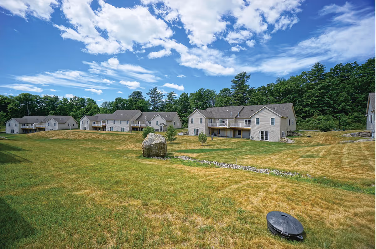 View of a senior living community named Black Rocks Village featuring multiple two-story beige residential buildings with balconies, surrounded by a large grassy area with a few trees and a rock, under a partly cloudy blue sky.