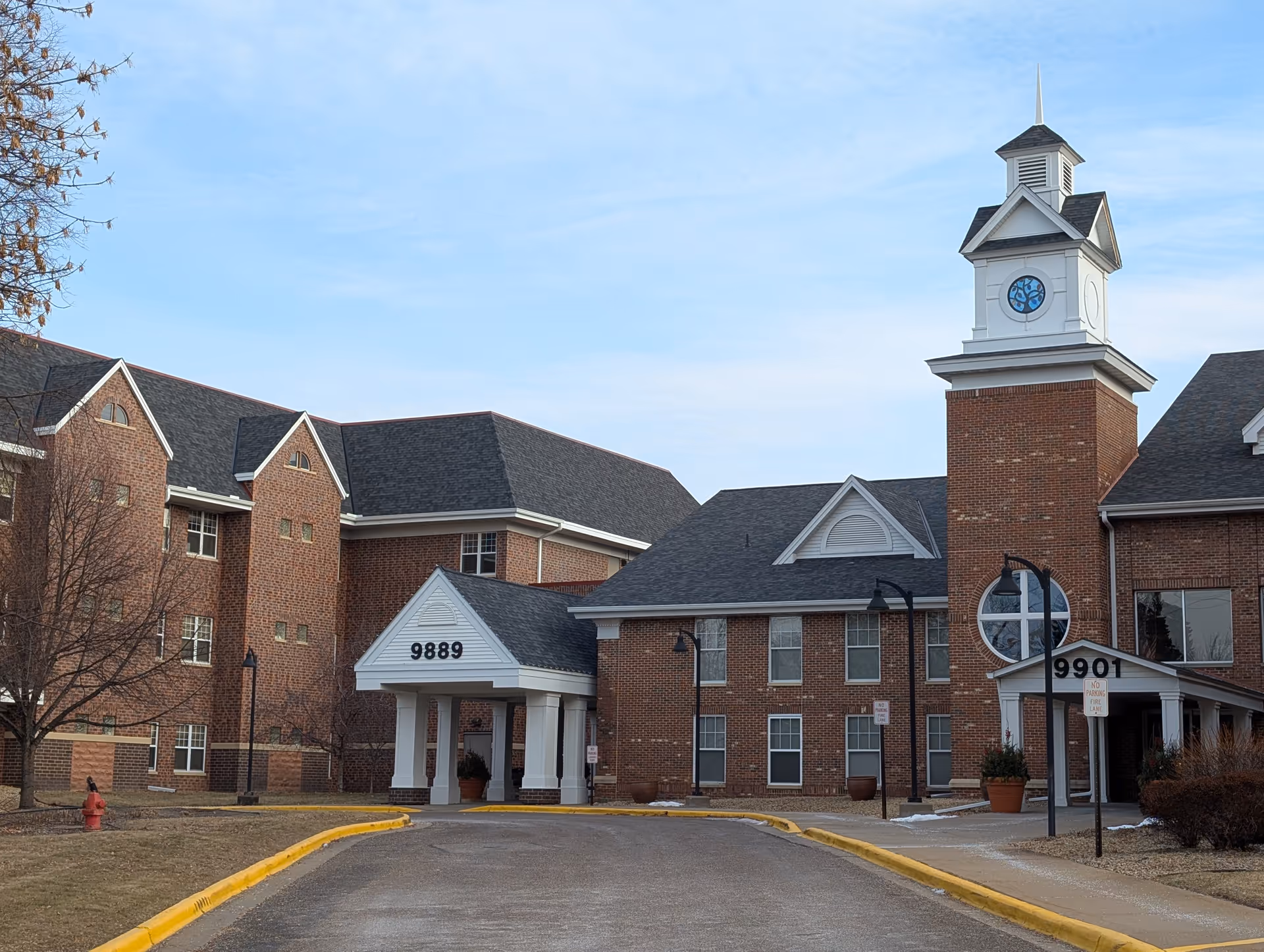 Front view of a brick senior living facility with a clock tower and two covered entrances labeled 9889 and 9901.