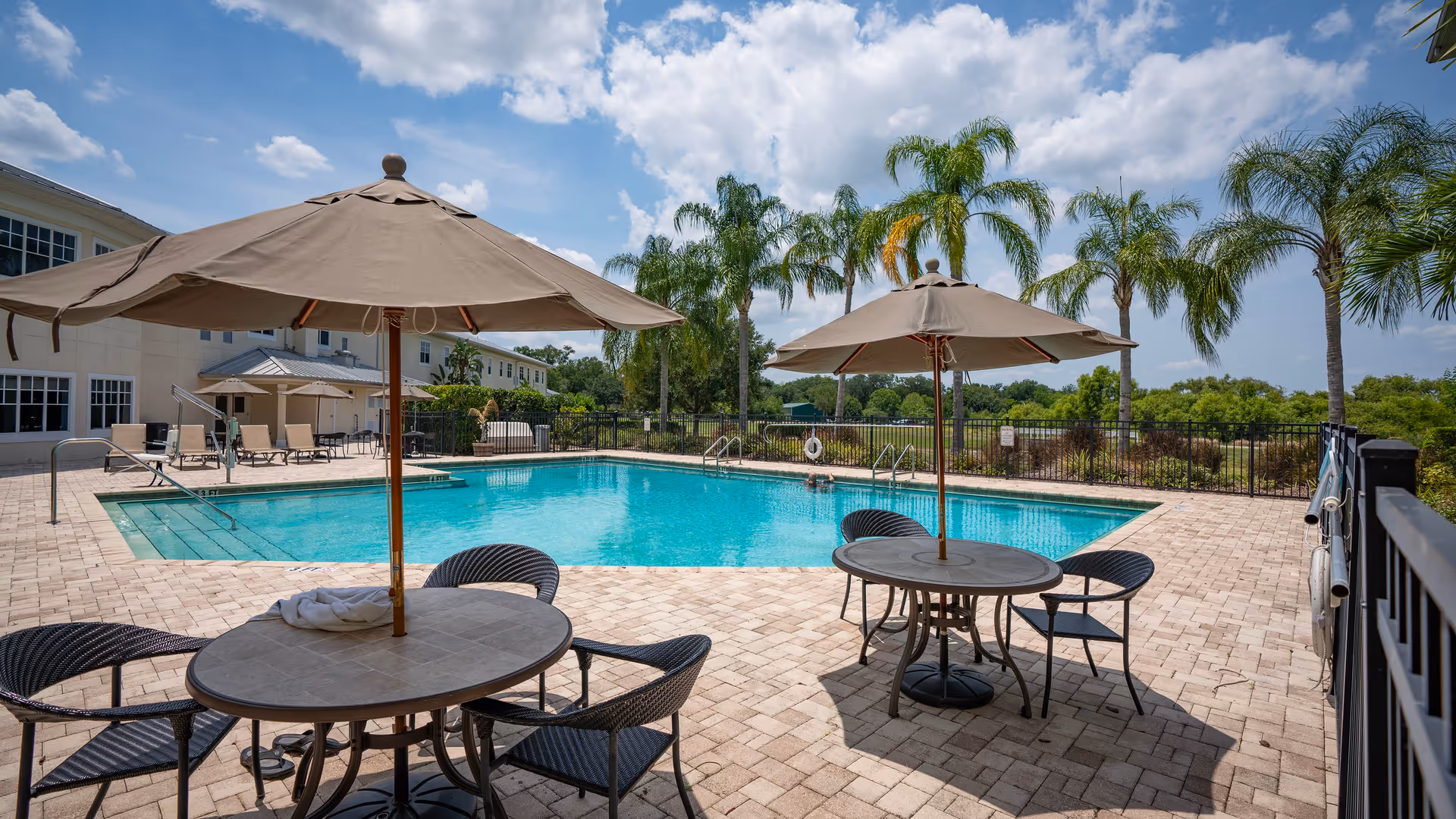 Outdoor swimming pool area with two round tables each shaded by large beige umbrellas and surrounded by black wicker chairs. The pool is fenced and palm trees are visible in the background under a partly cloudy blue sky. A building with multiple windows and lounge chairs is adjacent to the pool.