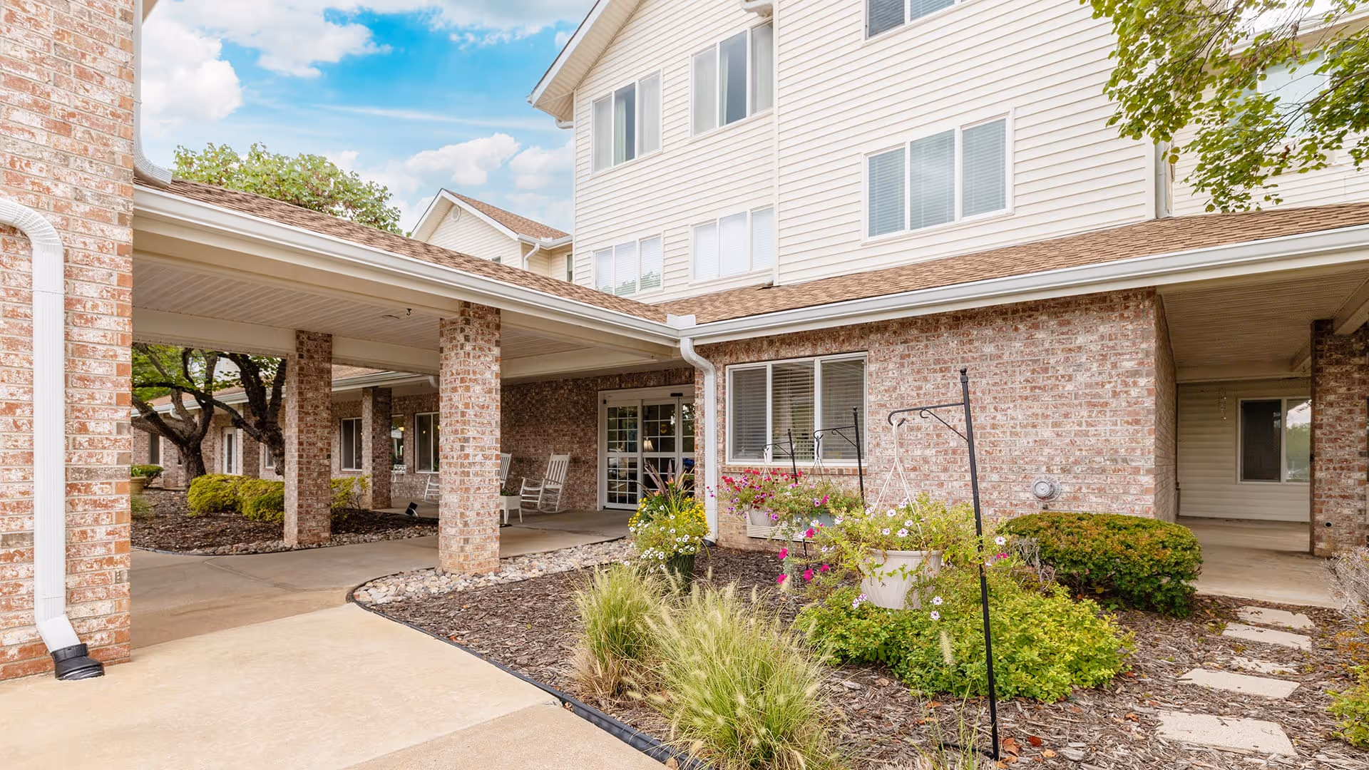 Exterior view of a senior living facility showing a covered walkway supported by brick pillars, landscaped garden beds with flowers and shrubs, and a multi-story building with brick and siding walls under a partly cloudy sky.