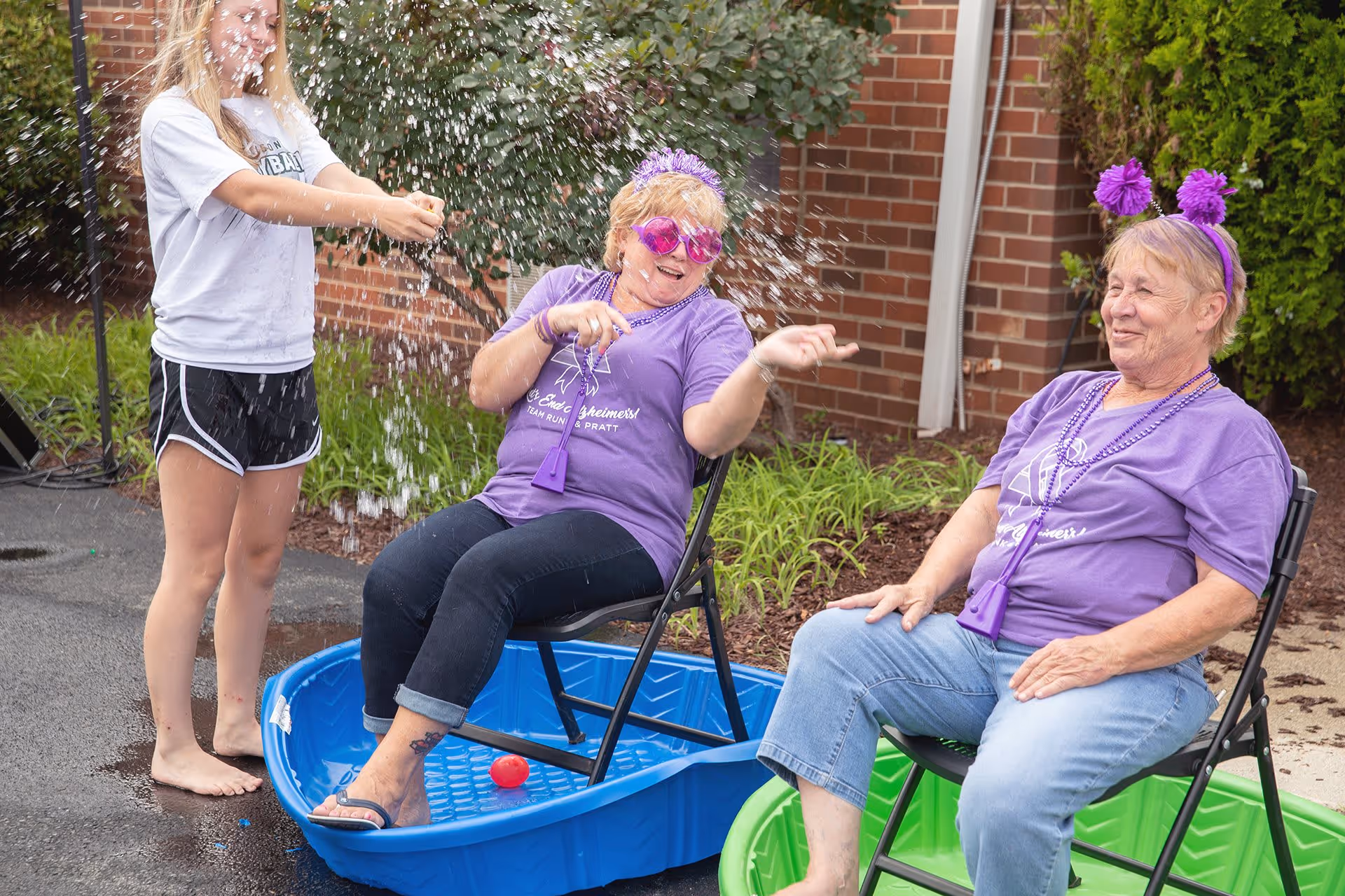 Two elderly women wearing purple shirts and festive headbands sit on chairs placed inside small plastic pools outdoors, while a younger person sprays water at them, creating a splash. They are smiling and enjoying the playful moment in front of a brick building with greenery around.