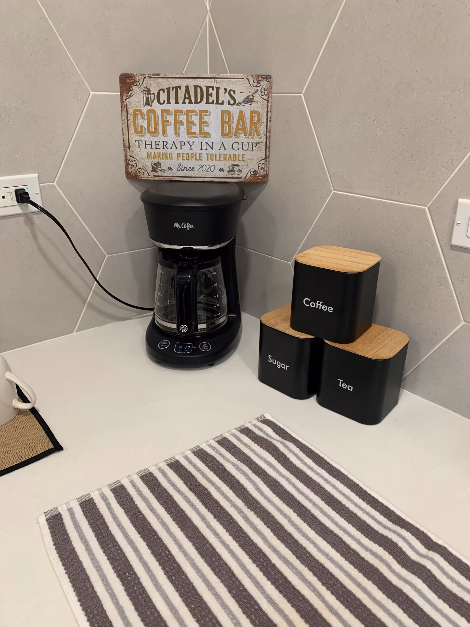 A coffee station with a black Mr. Coffee coffee maker, three black containers labeled Coffee, Sugar, and Tea with wooden lids, and a striped cloth on a white countertop. A decorative sign above the coffee maker reads 'Citadel's Coffee Bar Therapy in a Cup Making People Tolerable Since 2020'. The background features light gray hexagonal tiles.