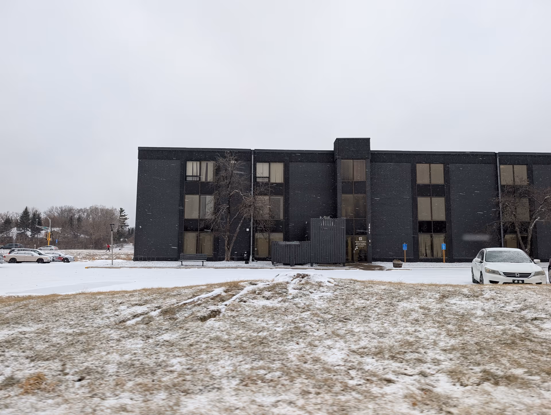 Snow-dusted lawn and parking lot in front of a three-story dark brick building under an overcast sky.