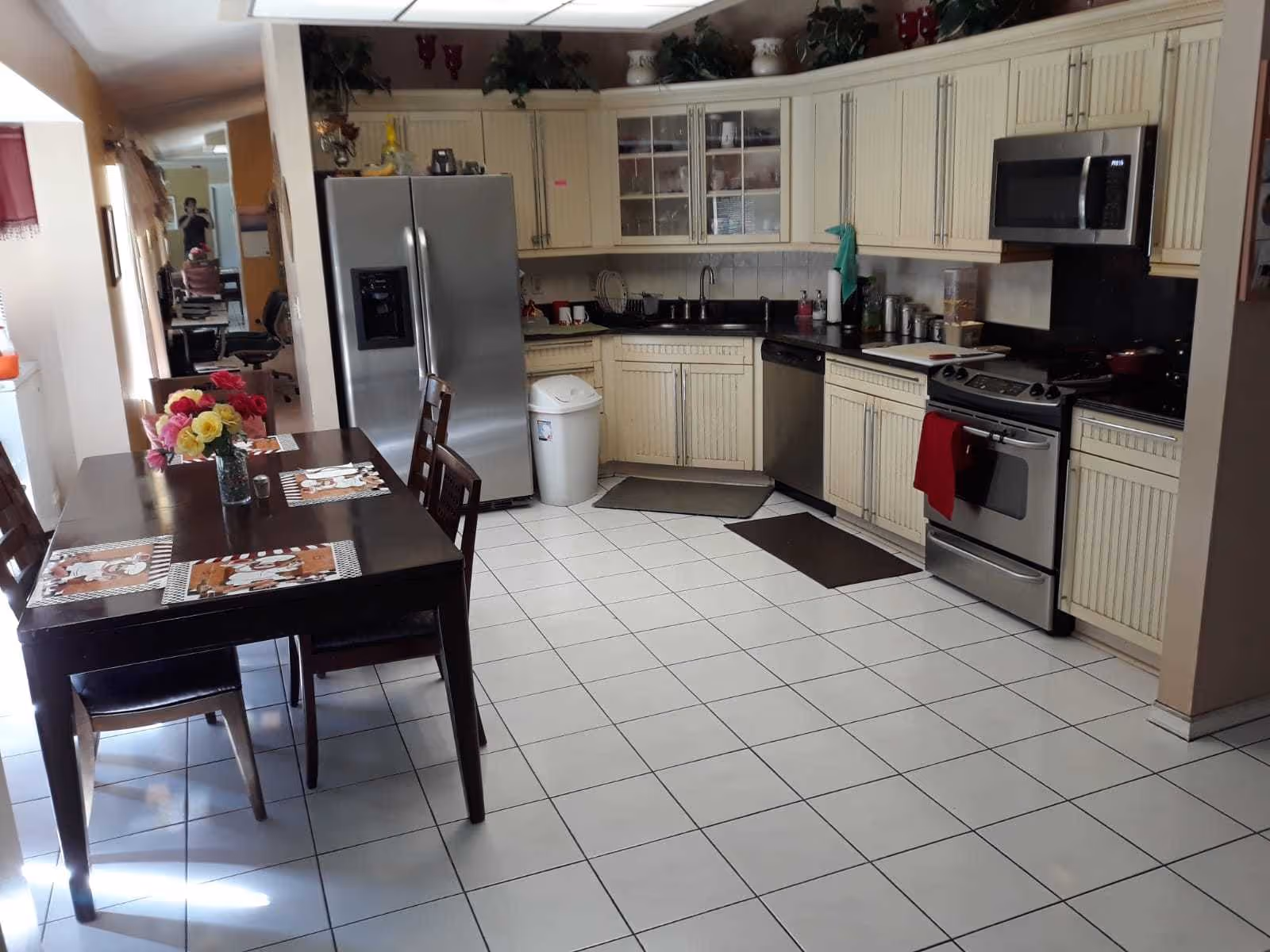 Bright combined kitchen and dining area with white tiled floor, cream cabinets, stainless steel refrigerator and stove, and a dark wood table with a vase of flowers.