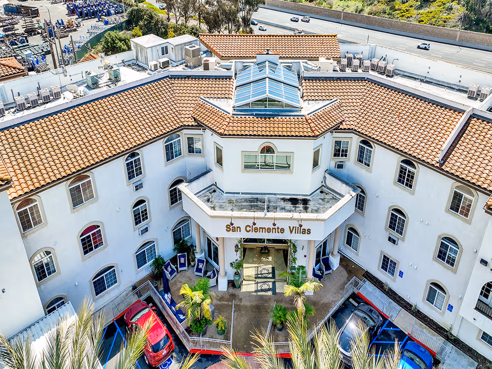 Aerial view of the front entrance of San Clemente Villas, a multi-story building with white walls and a red-tiled roof. The entrance features a covered porch with columns and the facility name displayed above. Several cars are parked in front, and palm trees and plants decorate the area. A highway is visible in the background.