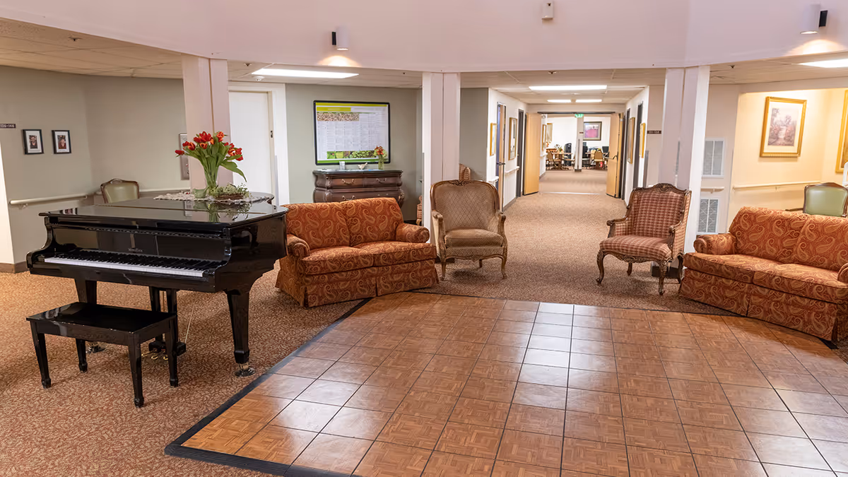 A senior living facility common area with a black grand piano, two patterned orange sofas, and two upholstered armchairs arranged around a square wooden dance floor. The space is carpeted with a hallway leading to other rooms in the background, decorated with framed pictures and soft lighting.