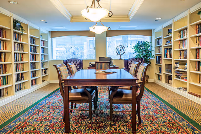 A well-lit library or reading room with a wooden table and six chairs in the center, surrounded by bookshelves filled with books on both sides. The room has large windows with blinds, a decorative rug under the table, and a ceiling light fixture. There is a globe and a potted plant near the windows.