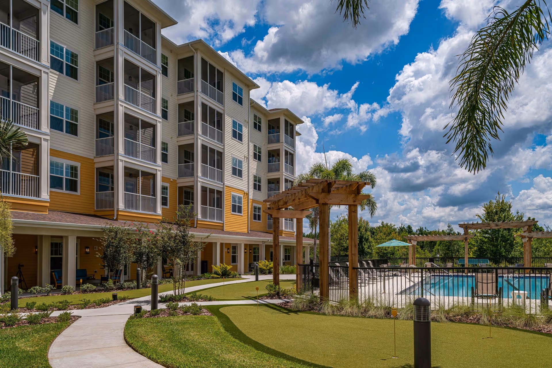 Outdoor view of Discovery Village Trinity showing a multi-story residential building with balconies, a putting green, palm trees, and a fenced swimming pool under a partly cloudy blue sky.