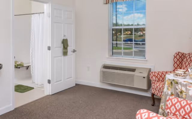 Small living area with patterned armchairs and a table, a wall air-conditioning unit under a window, and an open door revealing a bathroom.