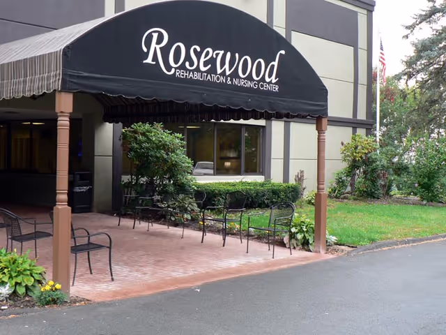 Entrance of Rosewood Rehabilitation & Nursing Center showing a black awning over a brick patio with outdoor chairs and landscaping.