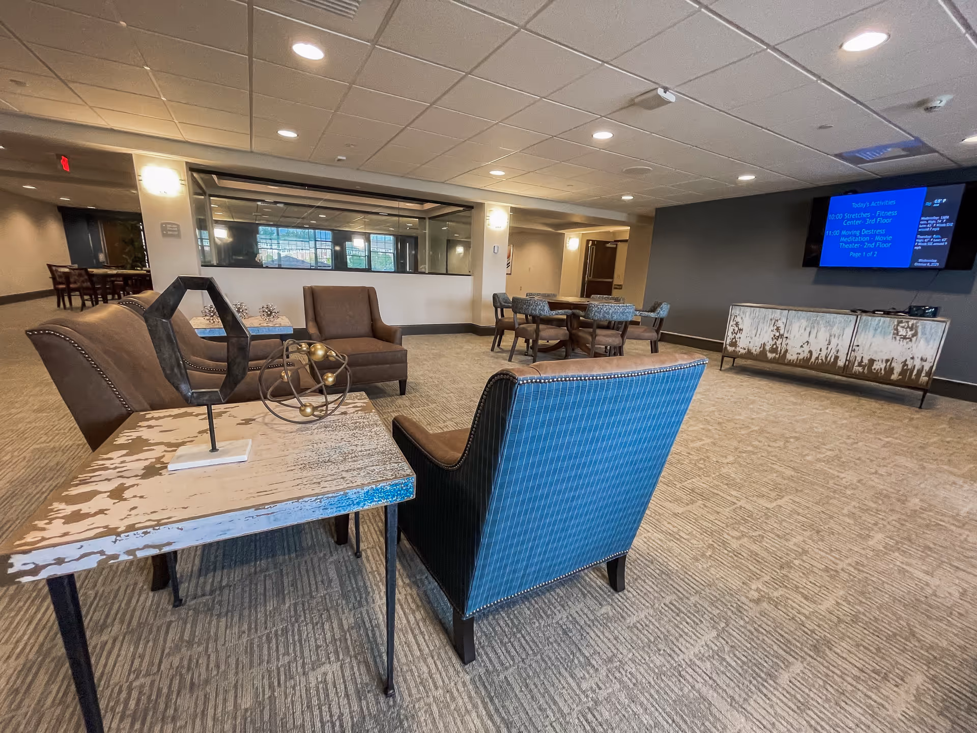 A spacious common area with various seating options including armchairs and a small table with decorative objects. In the background, there are additional tables and chairs, a large window, and a wall-mounted TV displaying today's activities. The room has carpeted flooring and a drop ceiling with recessed lighting.